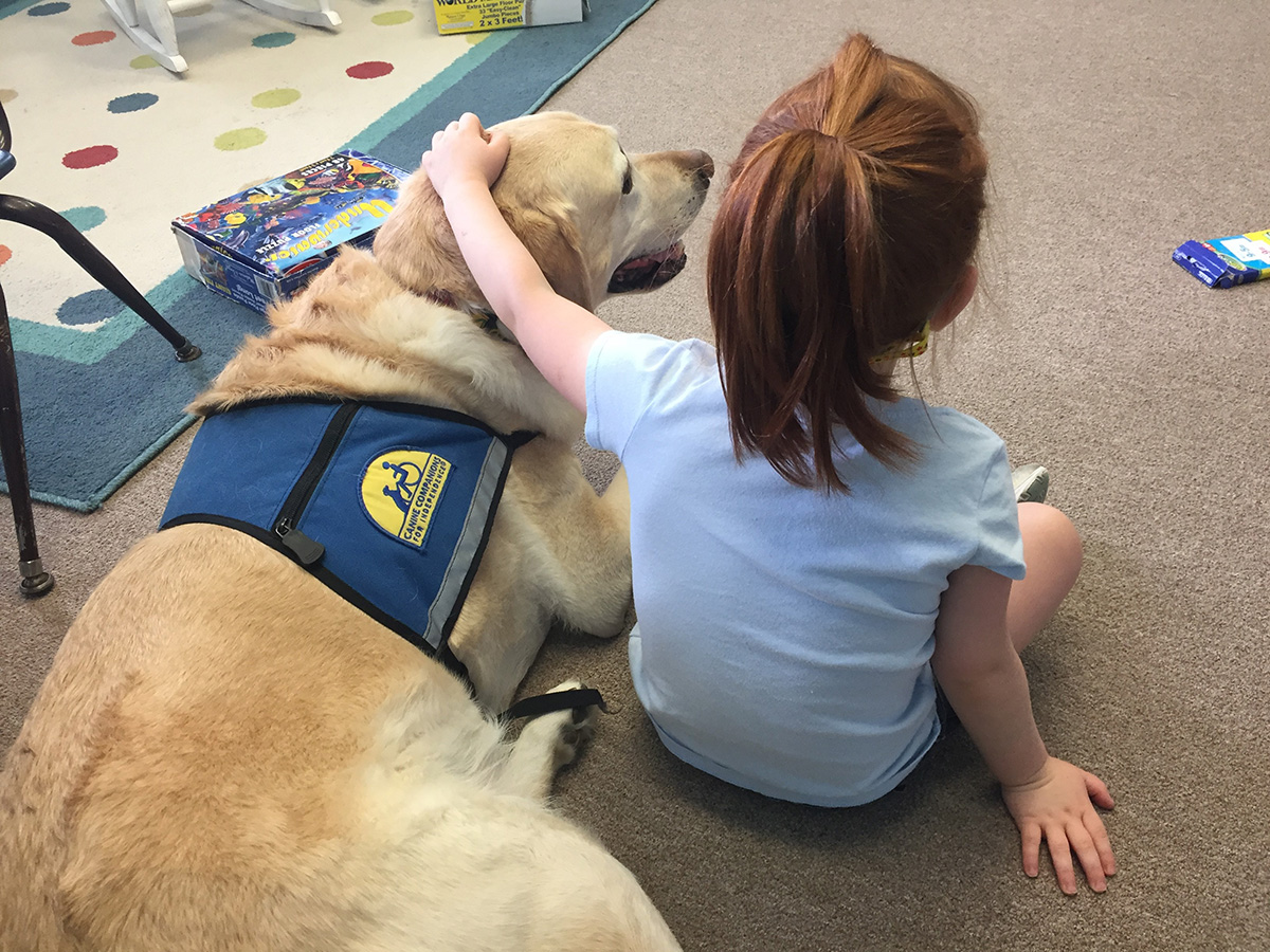 Pecos the facility dog lies on the carpet. A little red-headed girl sits with her arm on him, her hand on his head.