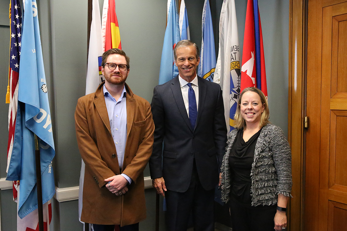 Nick Bratvold with Senator Thune and NCA's Denise Edwards in the senator's office, standing in front of flags.