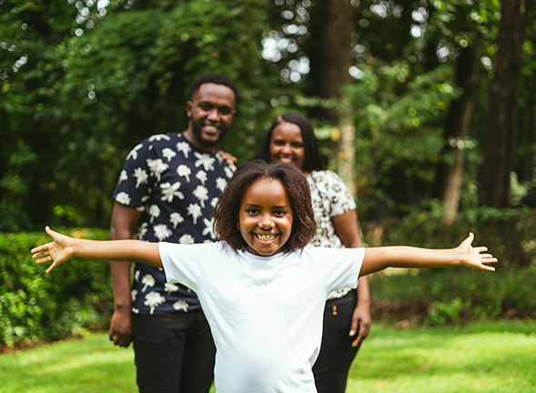 A young Black girl with her arms spread wide stands in front of her smiling parents. Photo by Osarugue Igbinoba from Unsplash.