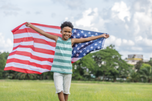 Black young boy streams an American flag over his shoulders in a field