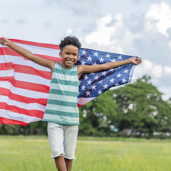 Black young boy streams an American flag over his shoulders in a field