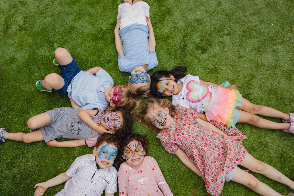Header image for June 3, 2025 blog from National Children's Alliance titled "The 10 Signs of Child Abuse You Need to Know." The image has 7 children laying down on the grass, smiling while looking at the camera. Their faces are painted. Stock image is from RDNE Stock Project uploaded to pexels.com