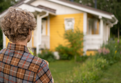 A teenage boy outside in a garden contemplatively stares at a small yellow country house where he lives