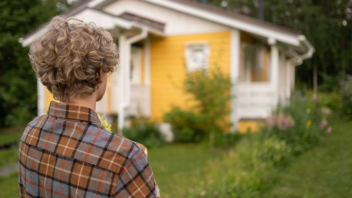 A teenage boy outside in a garden contemplatively stares at a small yellow country house where he lives